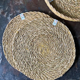 A top down view of the 6 Respiin seagrass tablemat in natural with a matching holder on top of a stone surface Features a brand patch stitched on the rim.