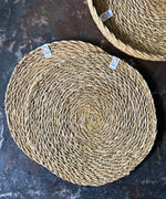 A top down view of the 6 Respiin seagrass tablemat in natural with a matching holder on top of a stone surface Features a brand patch stitched on the rim.