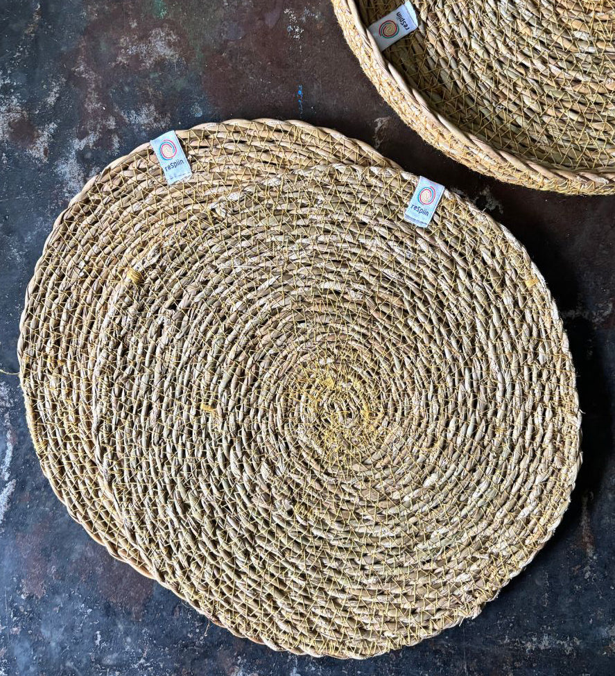 A top down view of the 6 Respiin seagrass tablemat in natural with a matching holder on top of a stone surface Features a brand patch stitched on the rim.