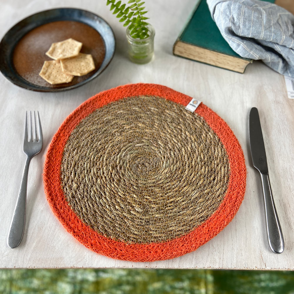 The ReSpiin seagrass and jute natural tablemat with an orange coloured ring, laid on a table.