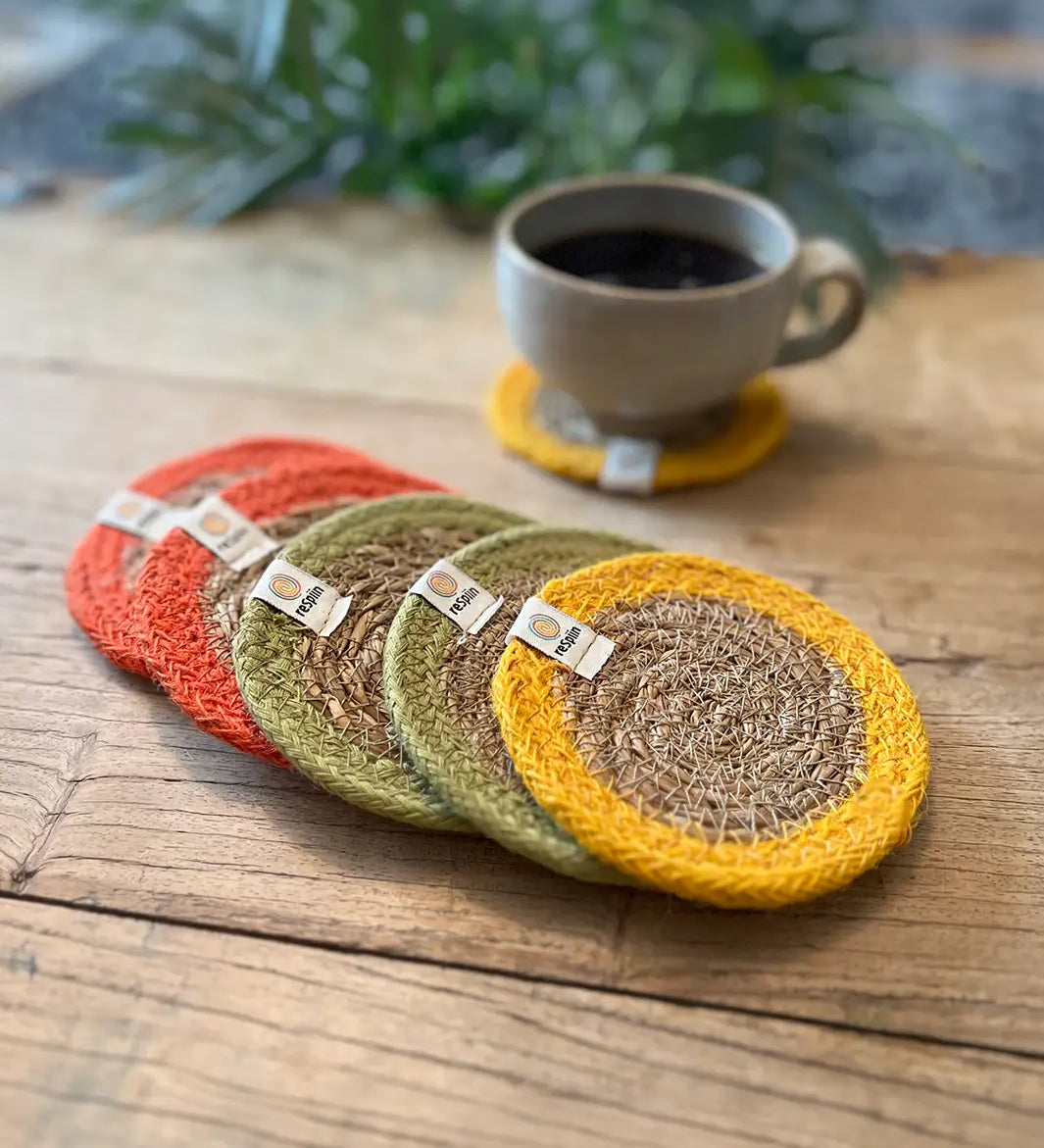 Set of 6 ReSpiin brights jute coasters with bright trim on a wooden table with a cup of coffee standing on one coaster