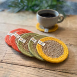 Set of 6 ReSpiin brights jute coasters with bright trim on a wooden table with a cup of coffee standing on one coaster