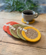 Set of 6 ReSpiin brights jute coasters with bright trim on a wooden table with a cup of coffee standing on one coaster
