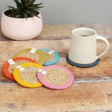 Set of 6 ReSpiin rainbow jute coasters on  a wooden table with a cup of coffee standing on one coaster