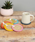 Set of 6 ReSpiin rainbow jute coasters on  a wooden table with a cup of coffee standing on one coaster