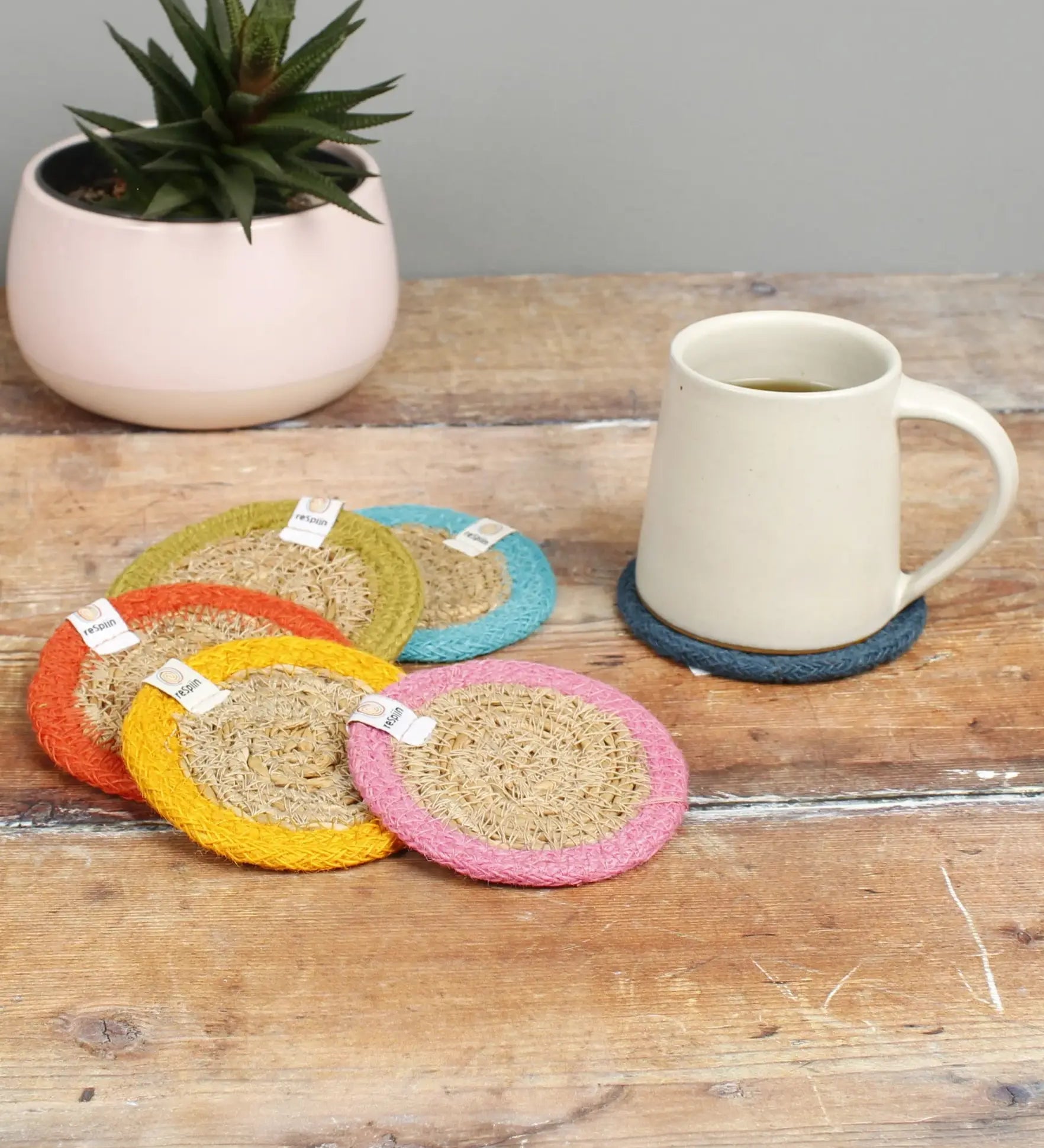 Set of 6 ReSpiin rainbow jute coasters on  a wooden table with a cup of coffee standing on one coaster