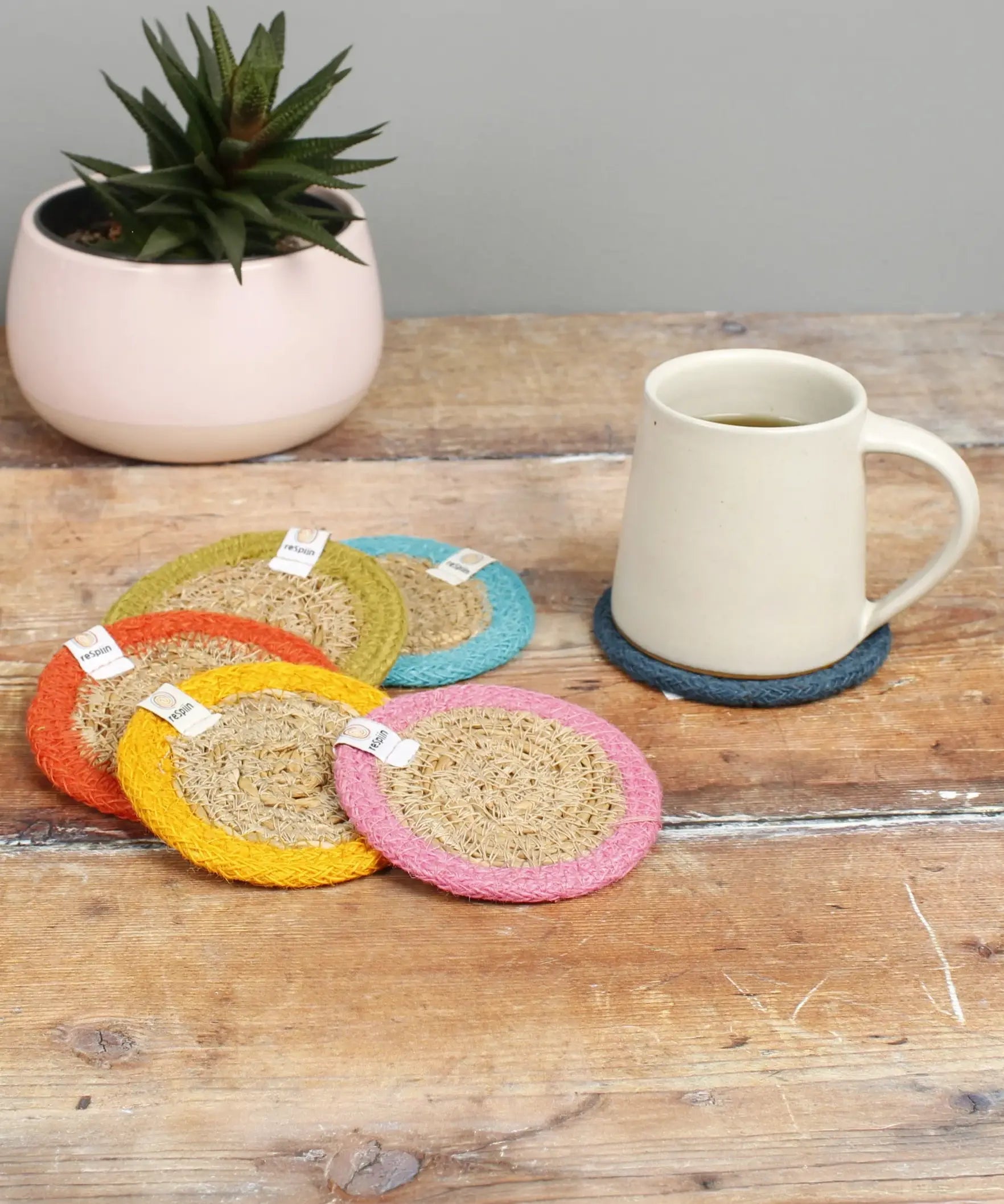 Set of 6 ReSpiin rainbow jute coasters on  a wooden table with a cup of coffee standing on one coaster