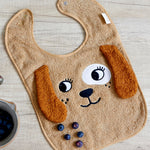The Roommate Dog Baby Bib with an embroidered puppy face with floppy 3D ears. On a tabletop with some blueberries. 