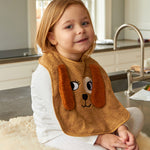 Child wearing the Roommate Dog Baby Bib with an embroidered puppy face with floppy 3D ears. She is sat on a worktop on a fleece rug.