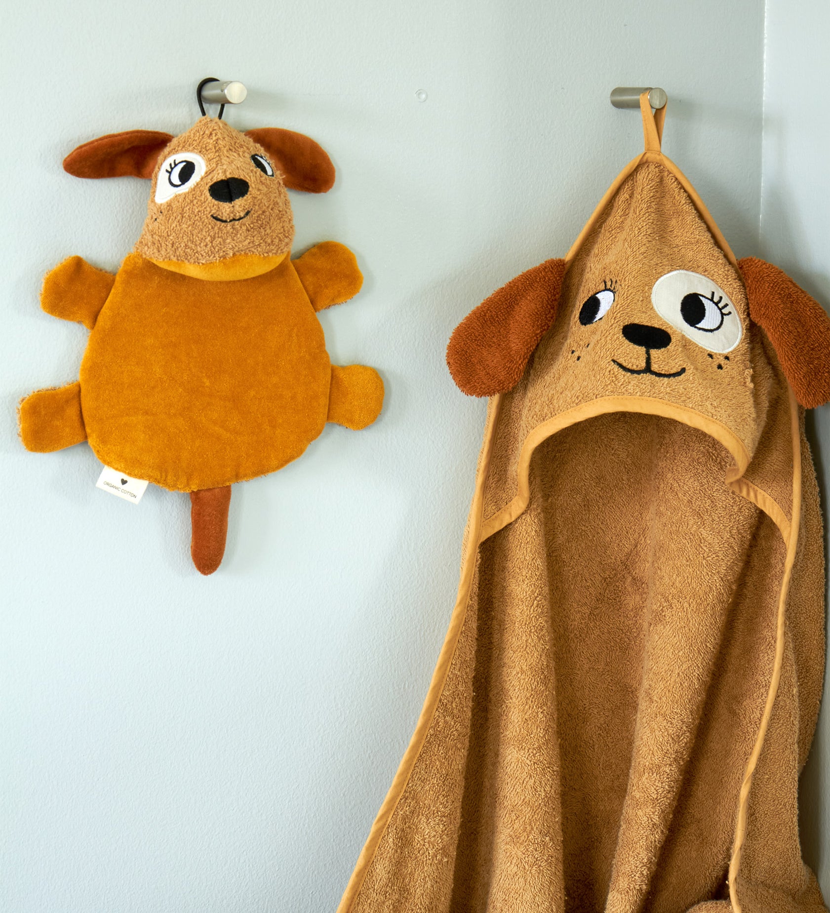 Roommate dog washcloth bath toy hanging on a wall hook next to a matching dog hooded towel, showing how the built-in loop is used for drying after bath time.