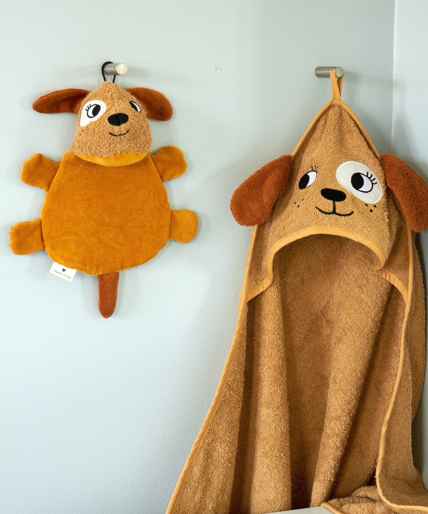 Roommate dog washcloth bath toy hanging on a wall hook next to a matching dog hooded towel, showing how the built-in loop is used for drying after bath time.
