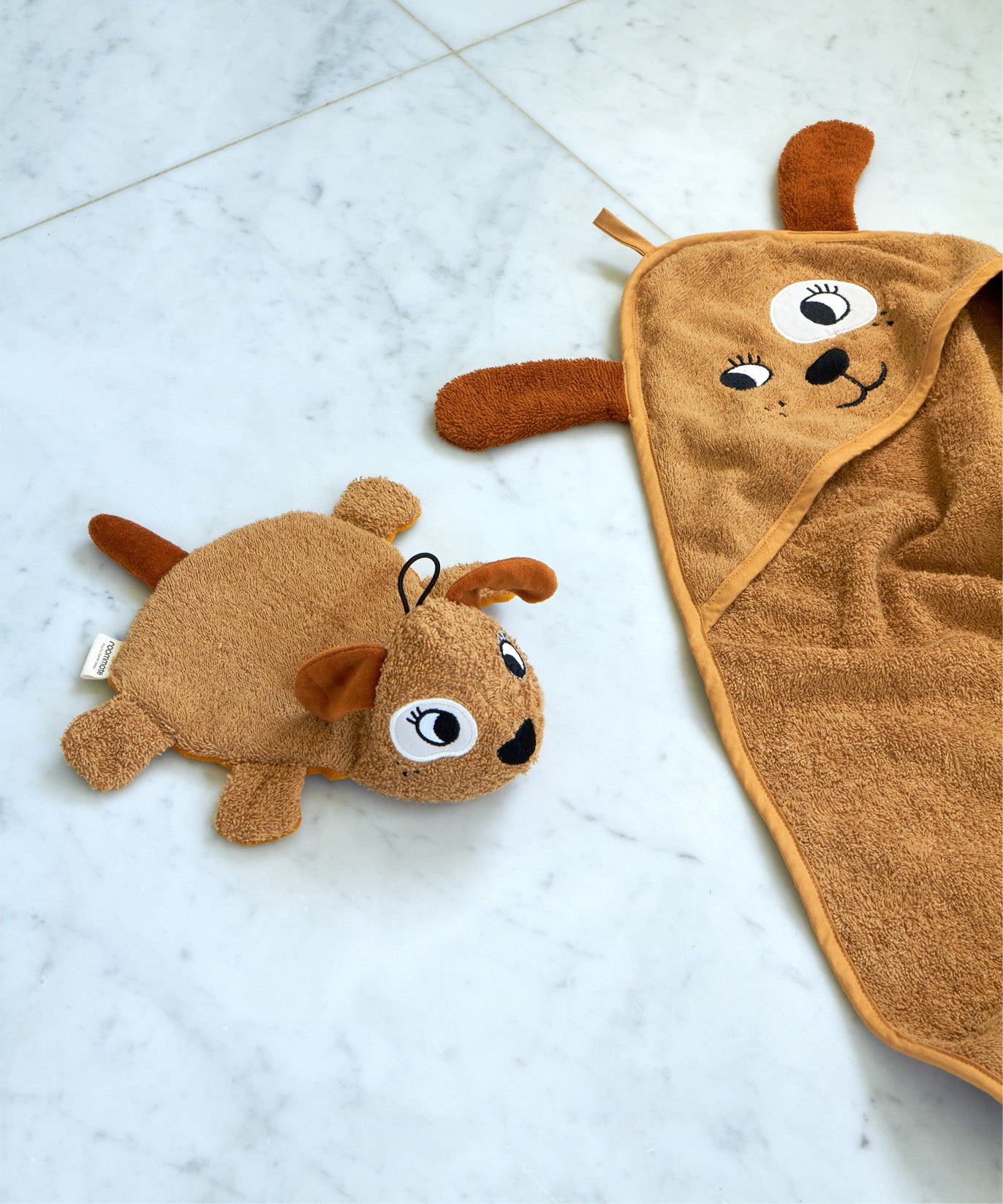 Roommate dog washcloth bath toy placed beside a matching dog hooded towel on a pale marble surface, showing coordinating organic cotton bathroom textiles.