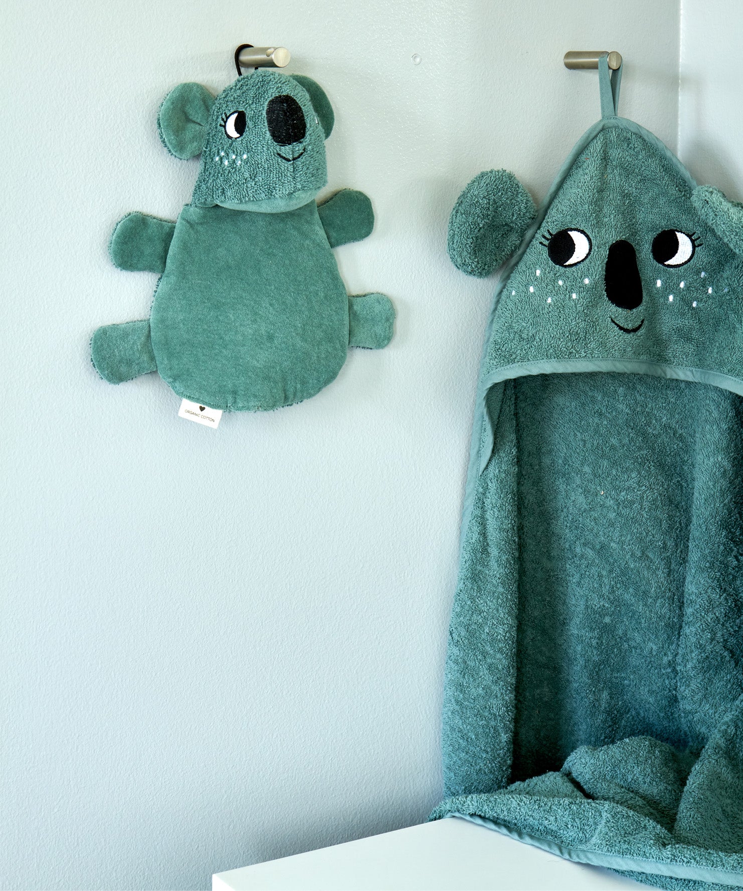Roommate koala washcloth bath toy hanging on a wall hook next to a matching koala hooded towel, showing the built-in hanging loop used for drying after bath time.