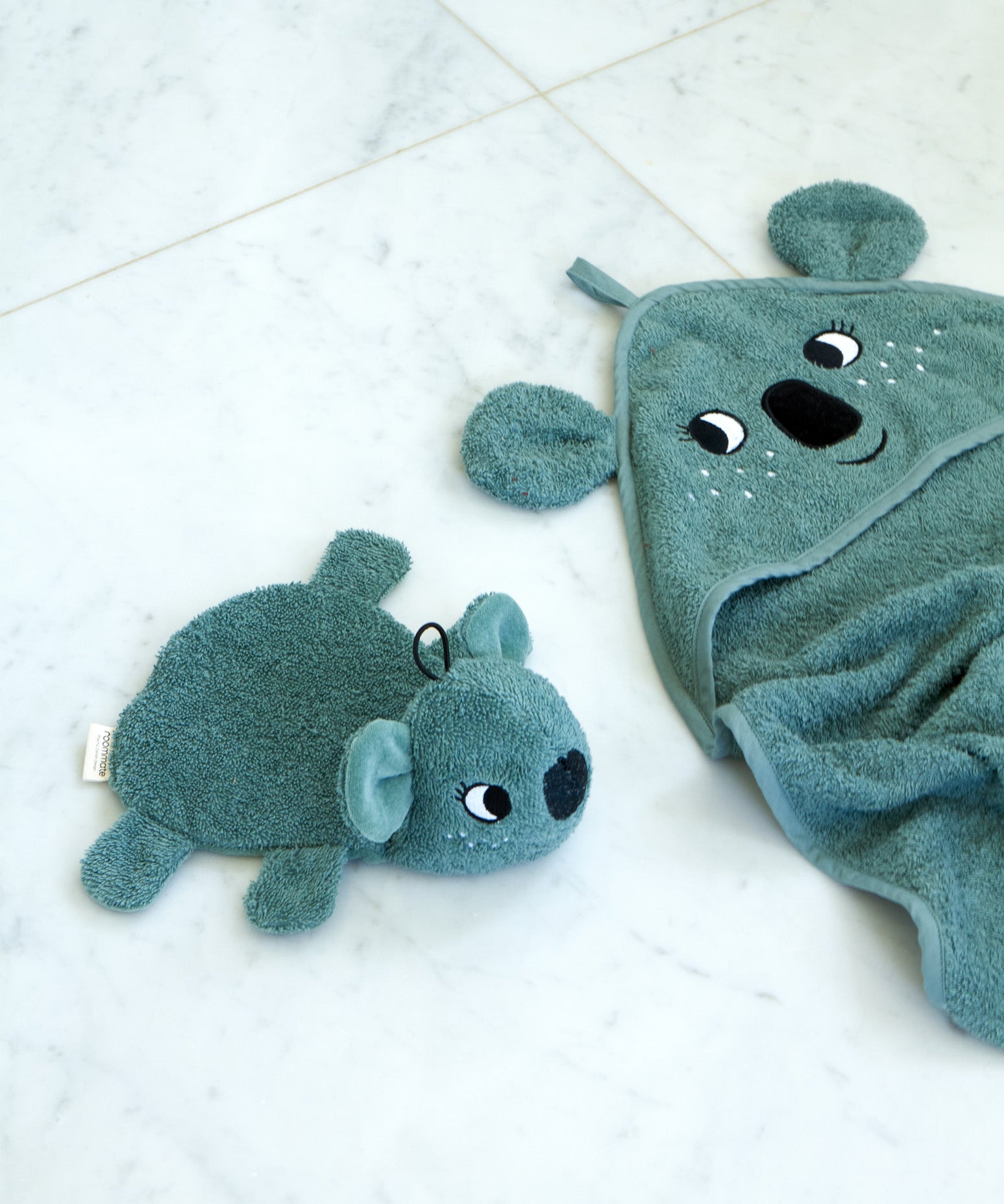 Roommate organic cotton koala washcloth bath toy placed beside a matching koala hooded towel on a pale surface, showing coordinating bathroom textiles from the Roommate range.