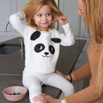Child sat on a grey kitchen worktop wearing the Roommate Panda Baby Bib in organic cotton with embroidered panda face with 3D ears. Parent is holding her and she has a bowl of blueberries.