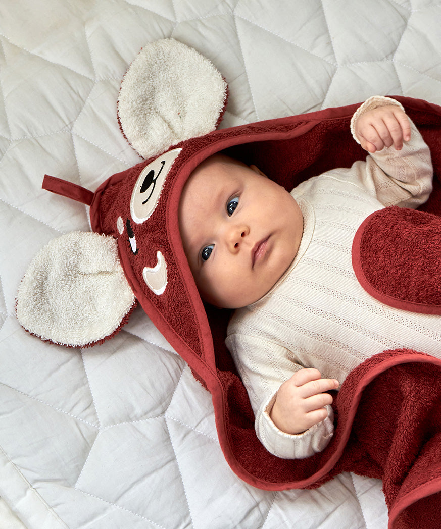 A little baby wrapped up in the Roommate organic cotton hooded towel, in red with a cute red panda face embroidered onto the hood. 