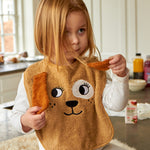 Child wearing the Roommate Dog Baby Bib with an embroidered puppy face with floppy 3D ears. She is sat on a worktop holding up the puppy ears.