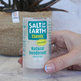 Adult hand holding a Salt Of The Earth classic unscented natural crystal deodorant stick showing scale with a plant in the background