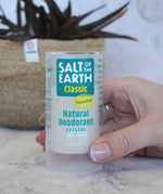 Adult hand holding a Salt Of The Earth classic unscented natural crystal deodorant stick showing scale with a plant in the background