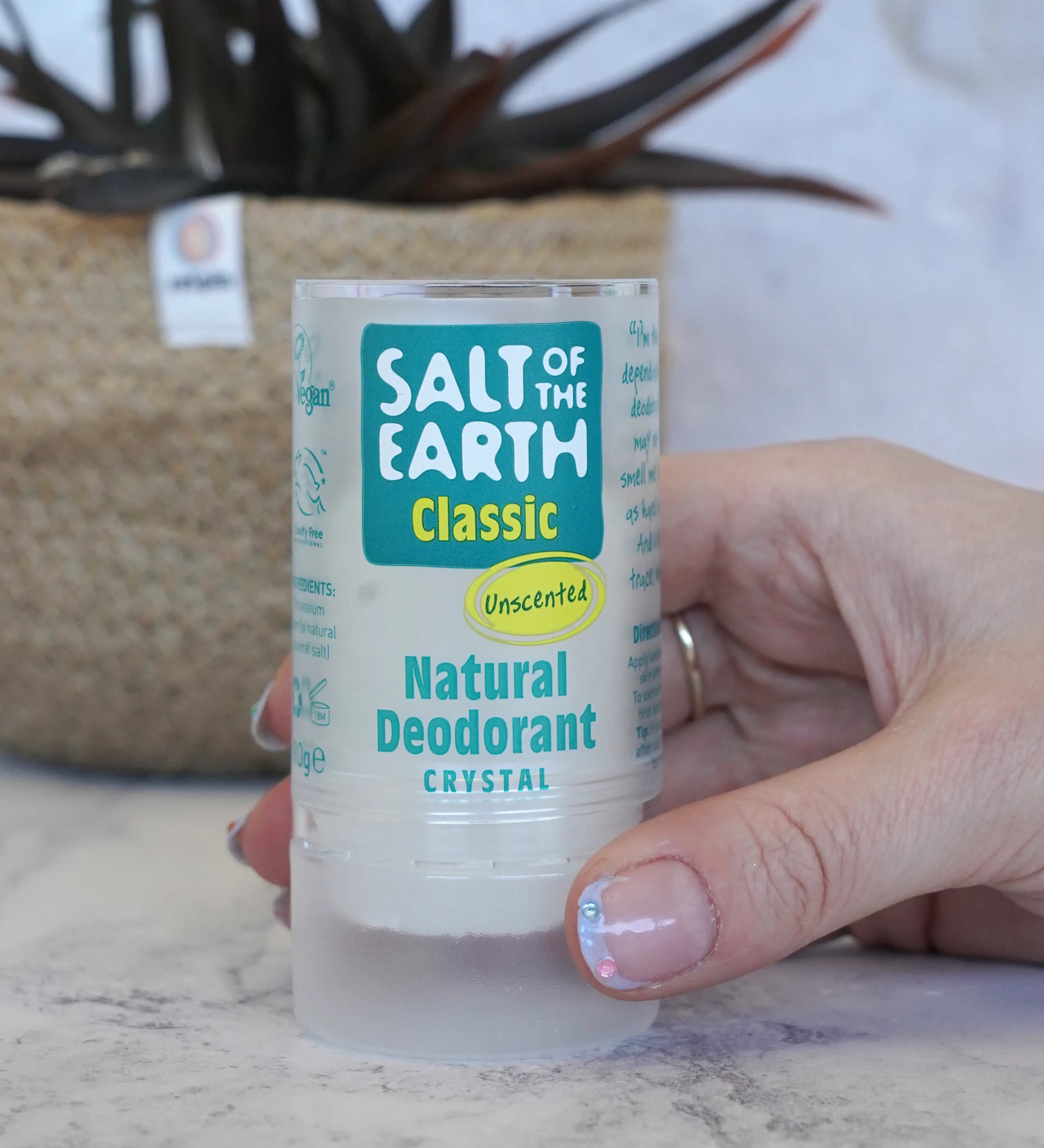 Adult hand holding a Salt Of The Earth classic unscented natural crystal deodorant stick showing scale with a plant in the background