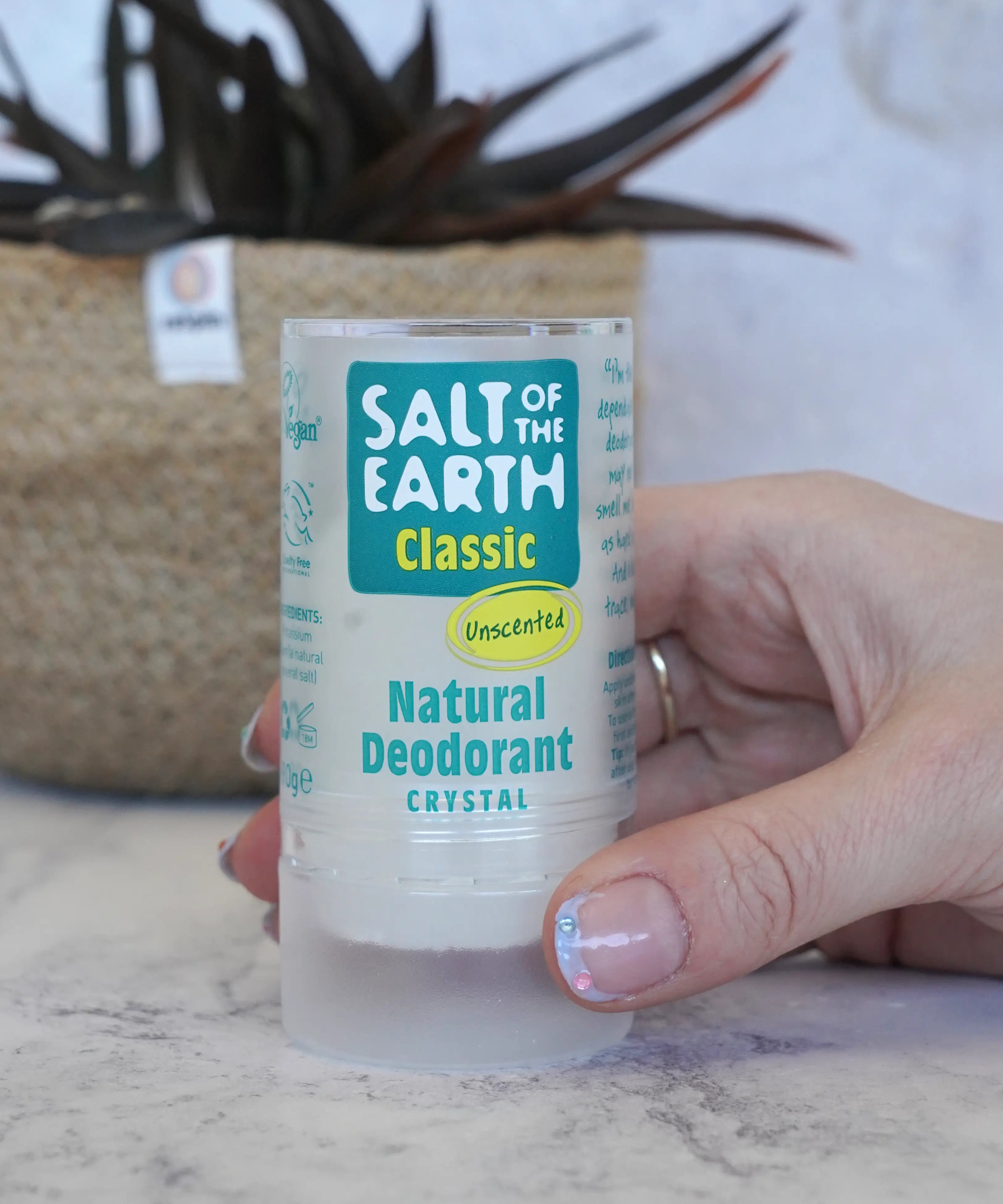 Adult hand holding a Salt Of The Earth classic unscented natural crystal deodorant stick showing scale with a plant in the background