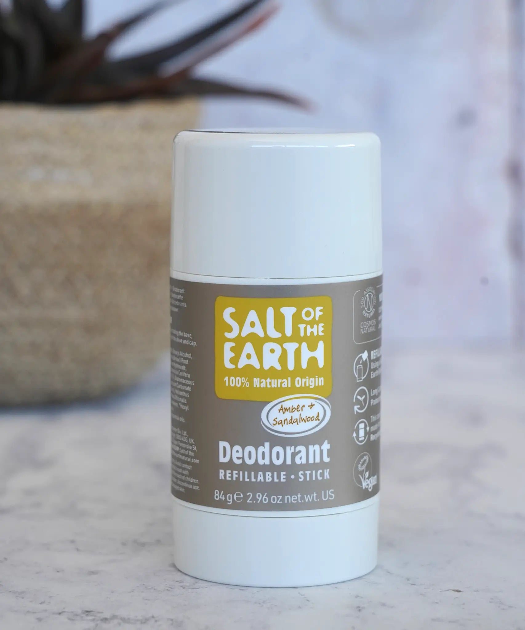 Salt Of The Earth refillable deodorant stick in natural amber and sandalwood on a marble surface with a plant in the background