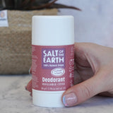 Adult hand holding the Salt Of The Earth refillable deodorant stick in lavender and vanilla showing scale with a plant in the background