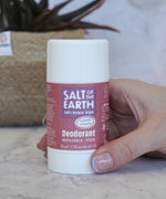 Adult hand holding the Salt Of The Earth refillable deodorant stick in lavender and vanilla showing scale with a plant in the background