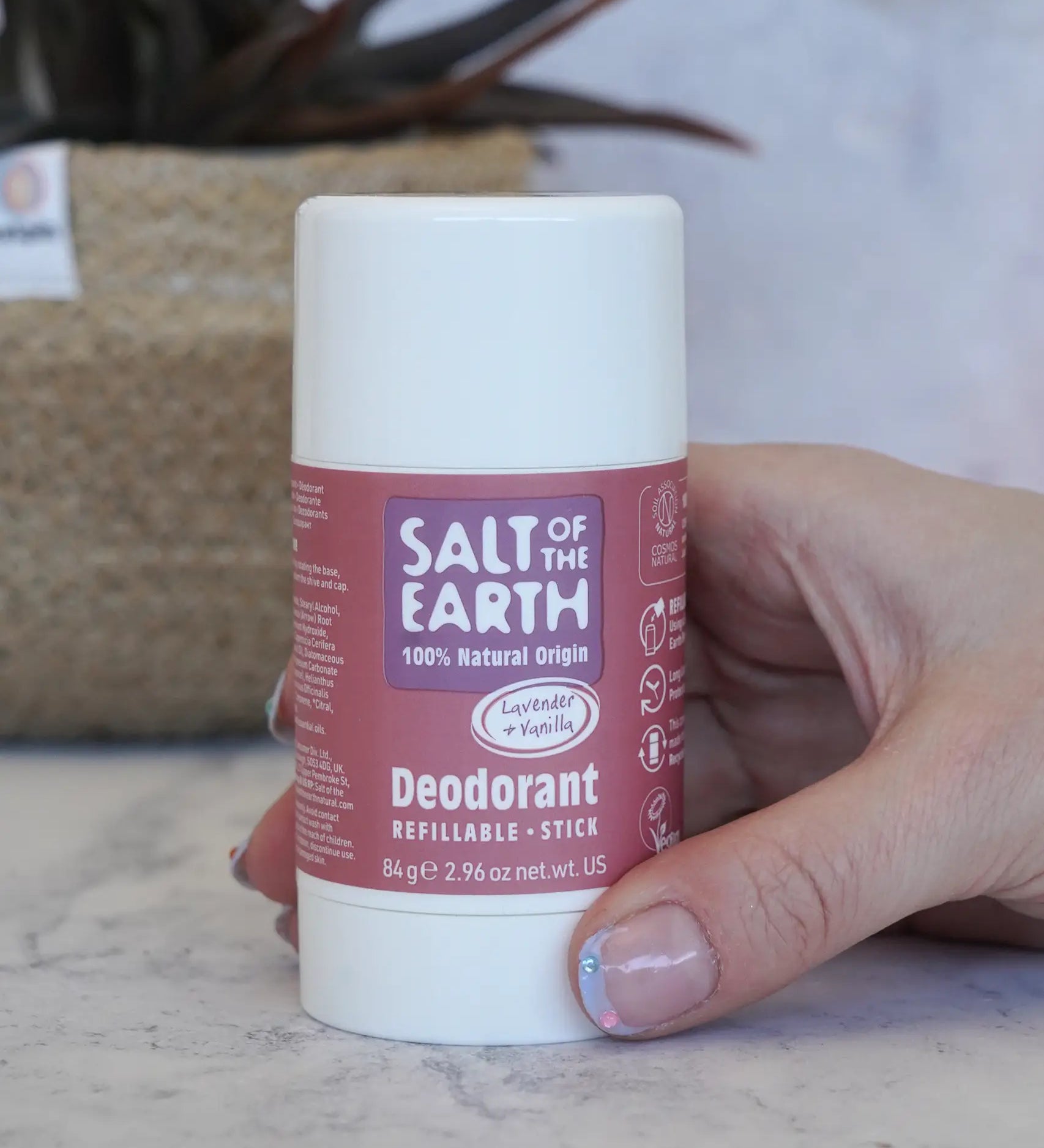 Adult hand holding the Salt Of The Earth refillable deodorant stick in lavender and vanilla showing scale with a plant in the background