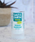 Salt Of The Earth classic unscented natural crystal deodorant stick standing on a marble surface with a plant in the background