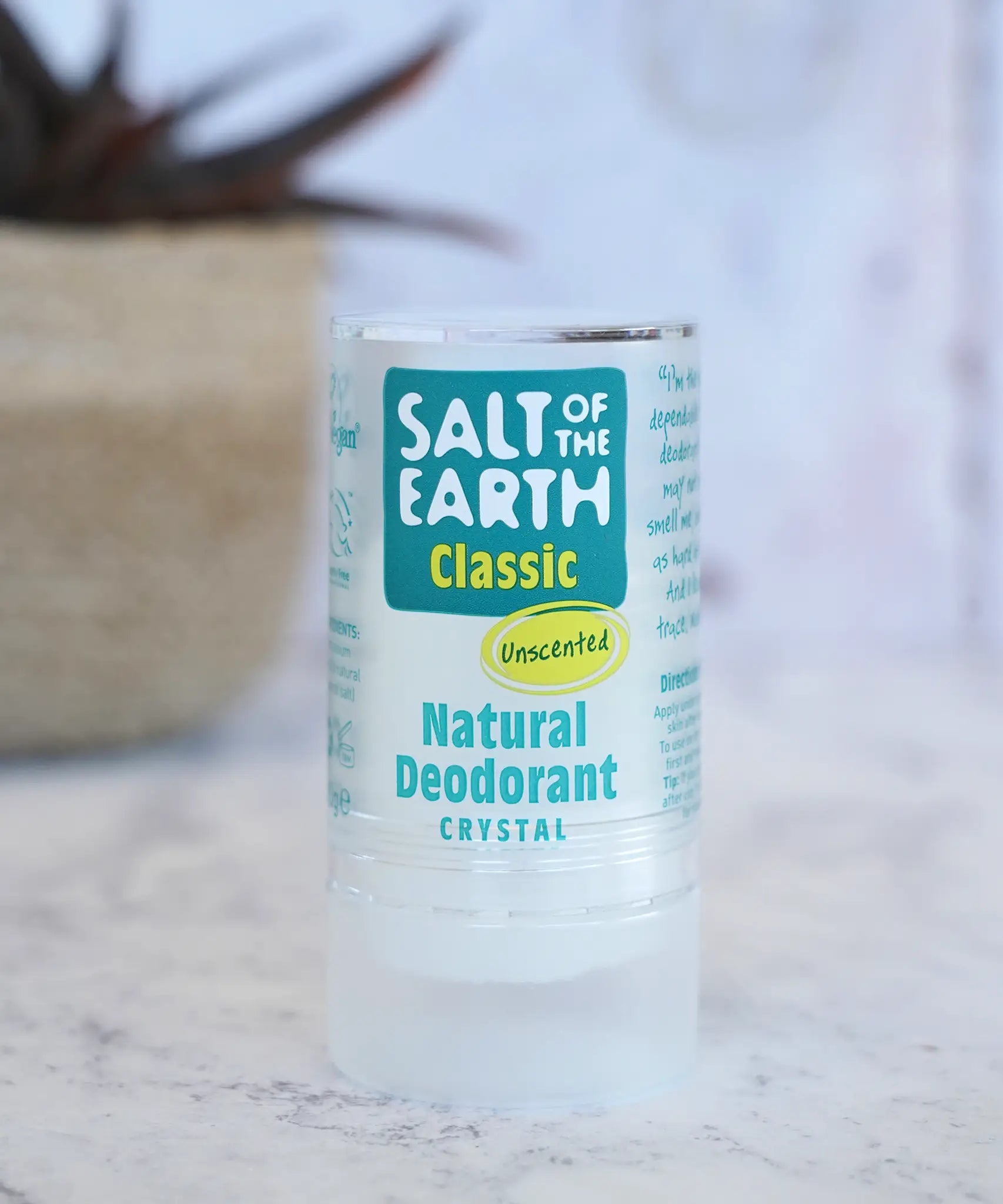 Salt Of The Earth classic unscented natural crystal deodorant stick standing on a marble surface with a plant in the background