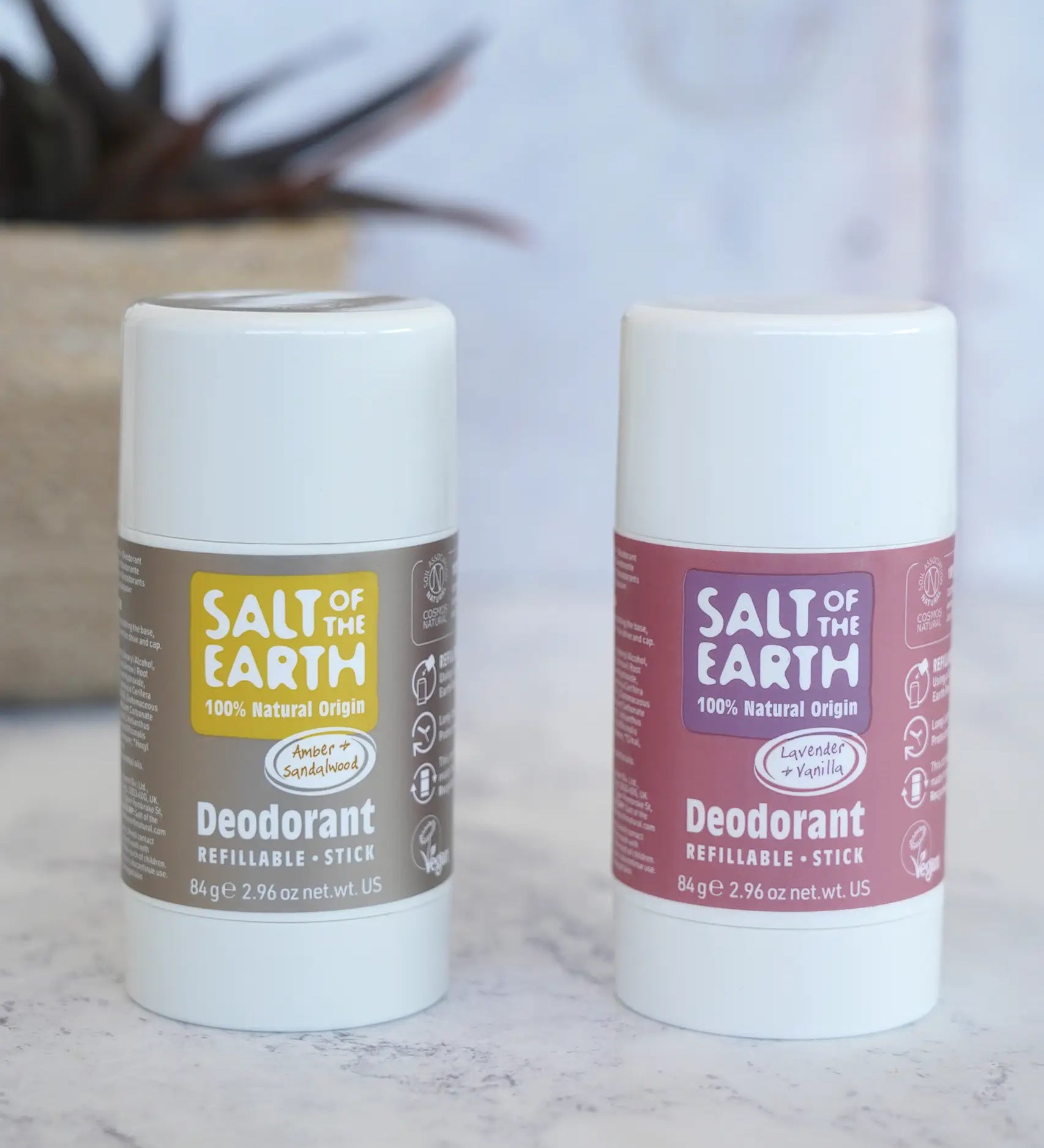Lavender and amber Salt Of The Earth refillable deodorant sticks standing on a marble surface with a plant in the background