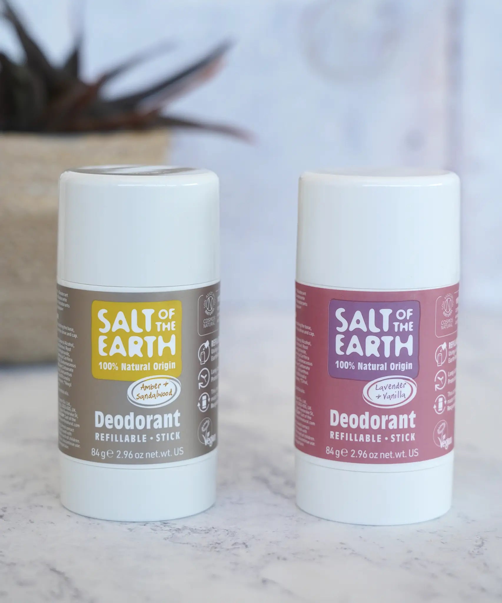 Lavender and amber Salt Of The Earth refillable deodorant sticks standing on a marble surface with a plant in the background