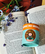 The Scrubber natural powder dry shampoo in 100% recycled cardboard packaging, in a ylang and rose scent on a open book next to blue flowers