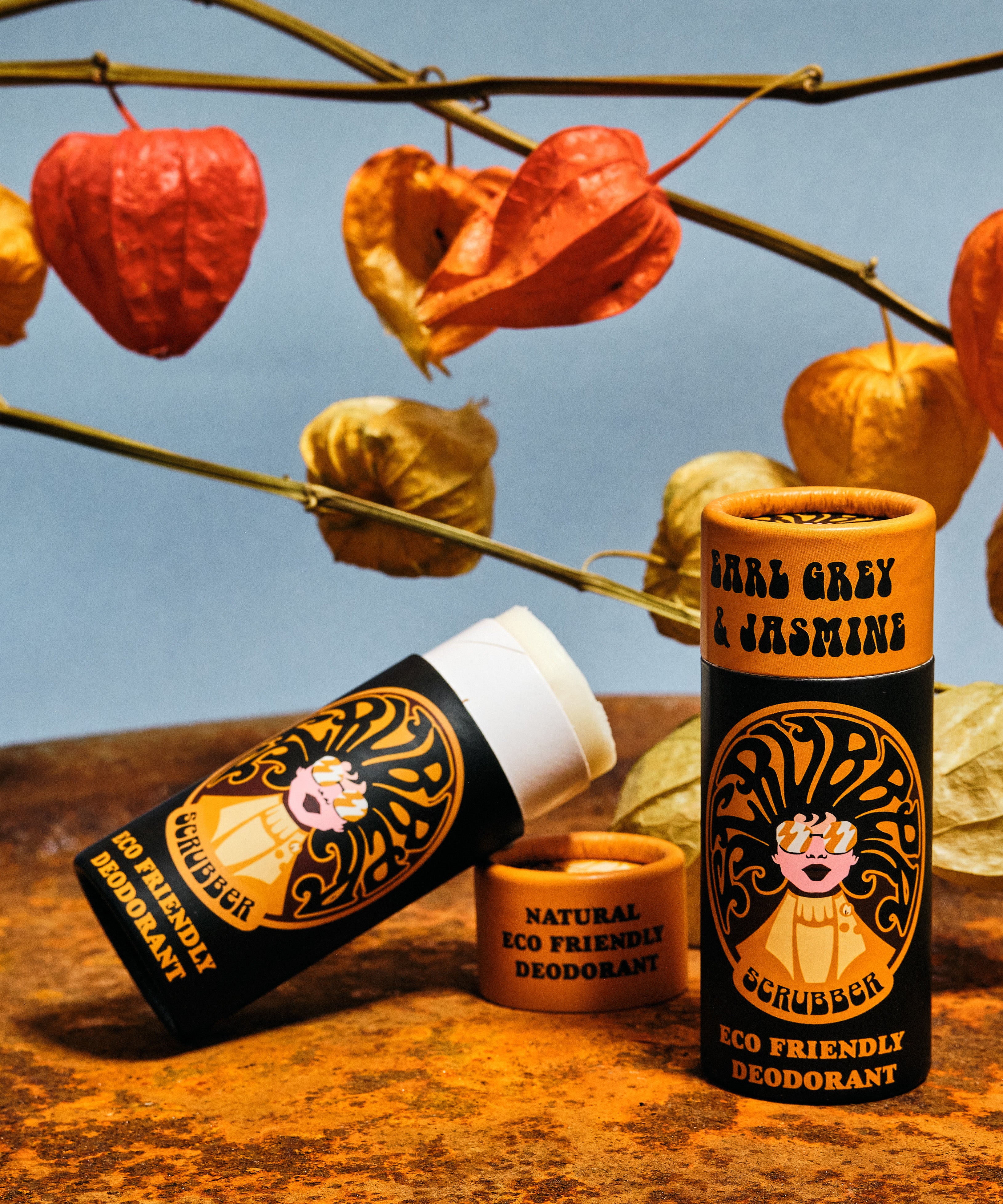 Two of The Scrubber plastic-free natural deodorant with groovy packaging standing on a metal surface under autumn coloured leaves