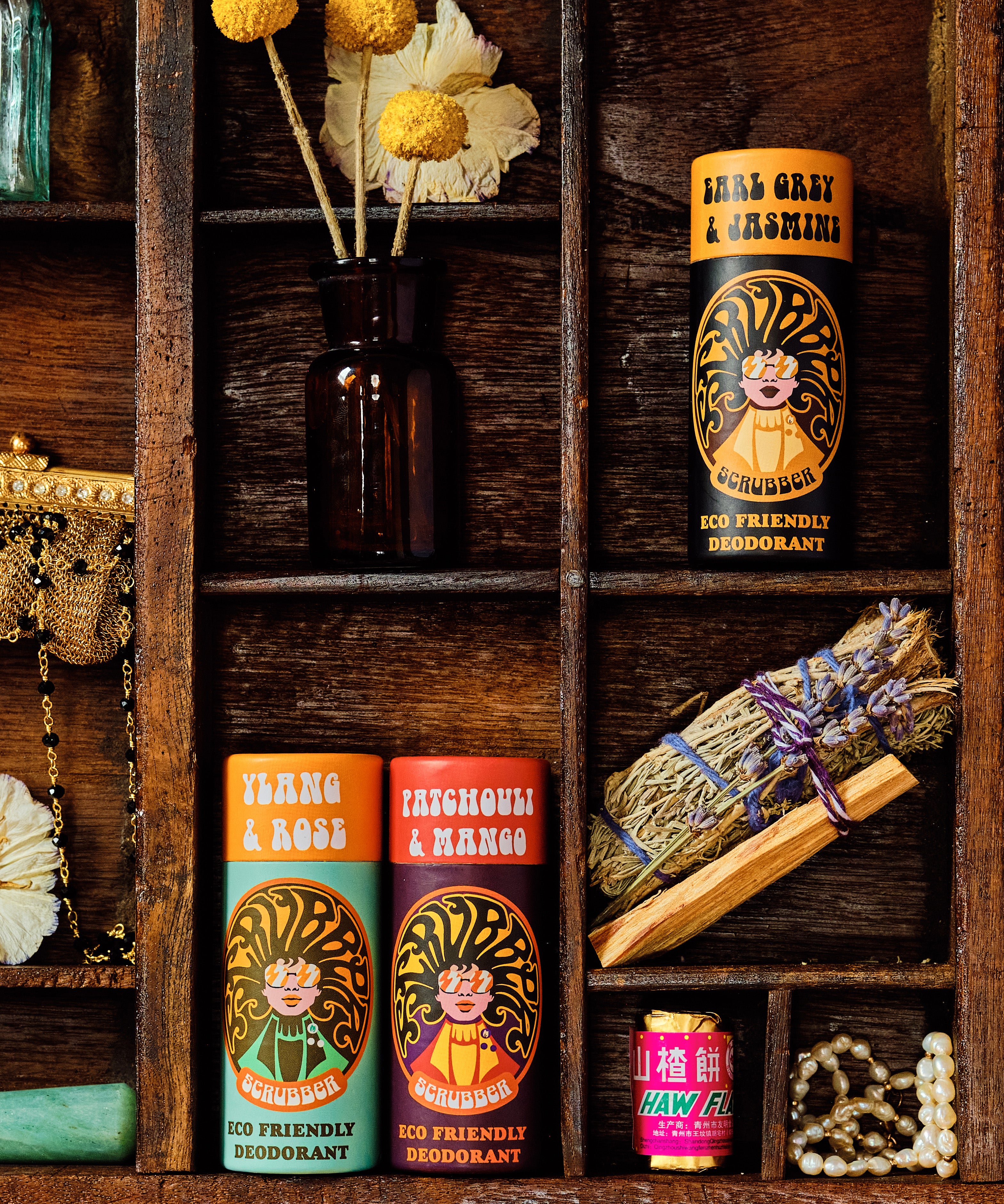 3 of The Scrubber plastic-free natural deodorant in ylang & rose, patchouli & mango and earl grey & jasmine scent with groovy packaging laid next to miscellaneous objects in a wooden display tray. 