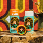 Two of The Scrubber plastic-free natural deodorant in ylang & rose with groovy packaging standing on a wooden surface with a psychedelic backdrop. 