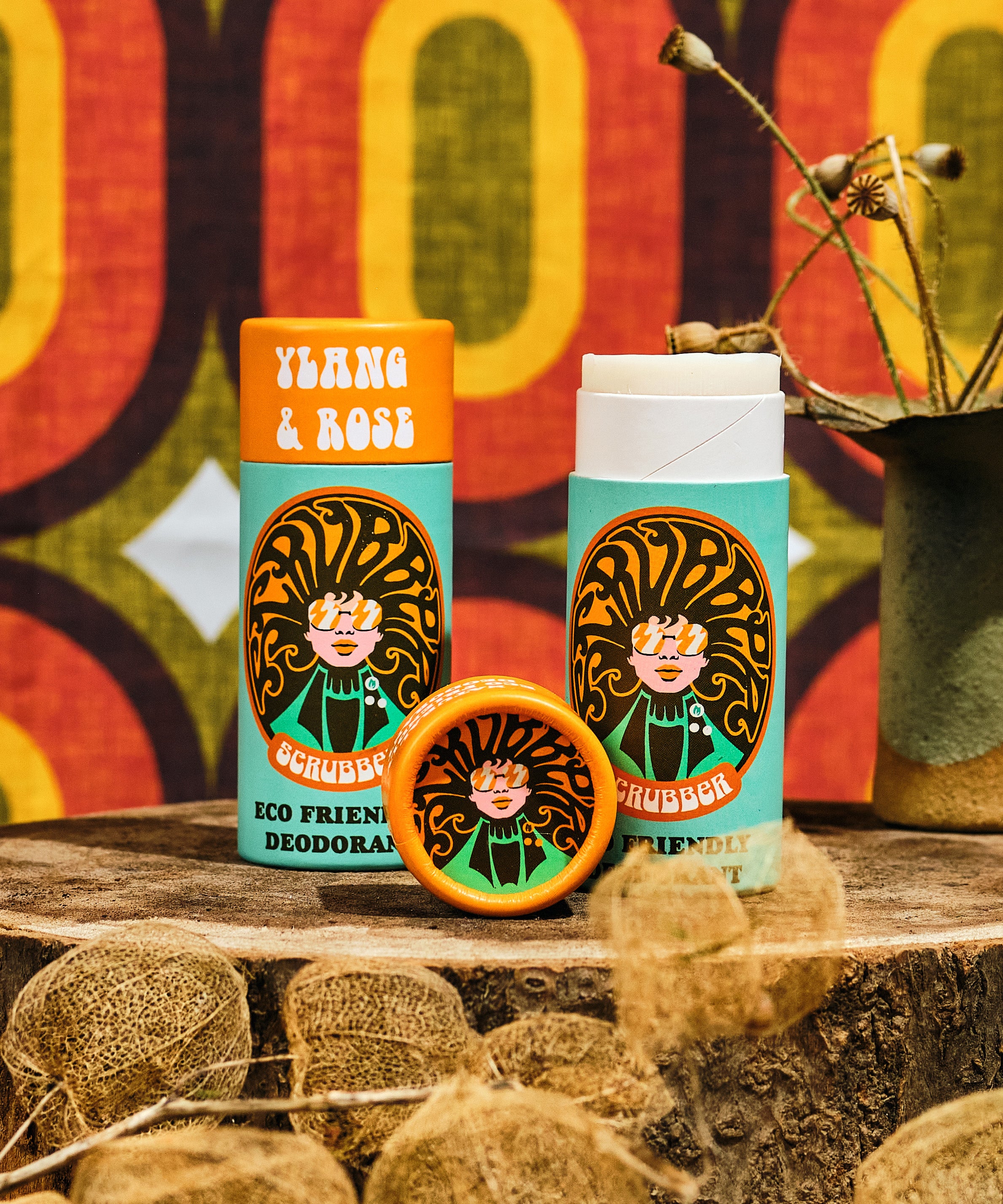 Two of The Scrubber plastic-free natural deodorant in ylang & rose with groovy packaging standing on a wooden surface with a psychedelic backdrop. 