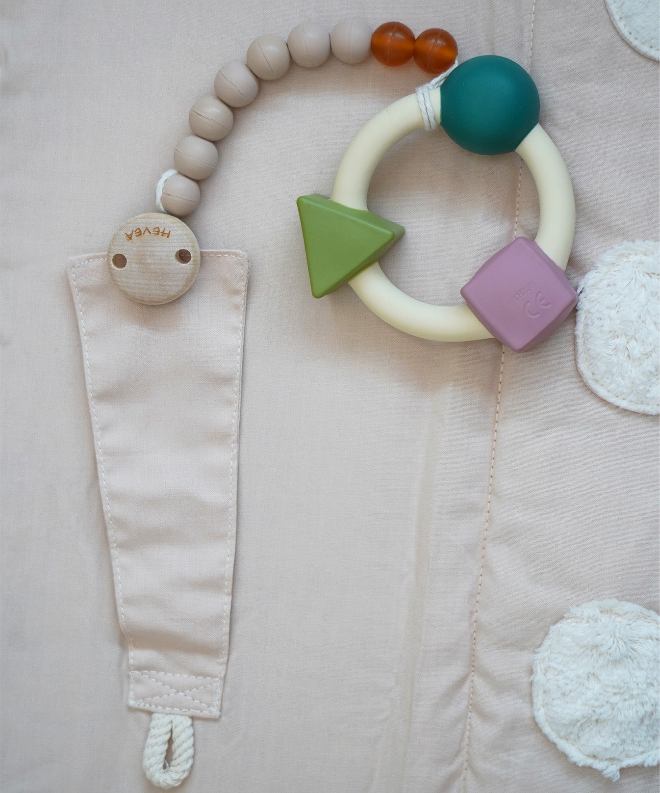 A natural rubber Hevea pacifier holder clip attached to the Senger Cozy Nap Playmat, with an Oli & Carol rubber teether attached.