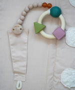 A natural rubber Hevea pacifier holder clip attached to the Senger Cozy Nap Playmat, with an Oli & Carol rubber teether attached.