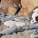 A collection of sea life themed cuddly animals from Senger placed on rocks and pebbles on a beach. 