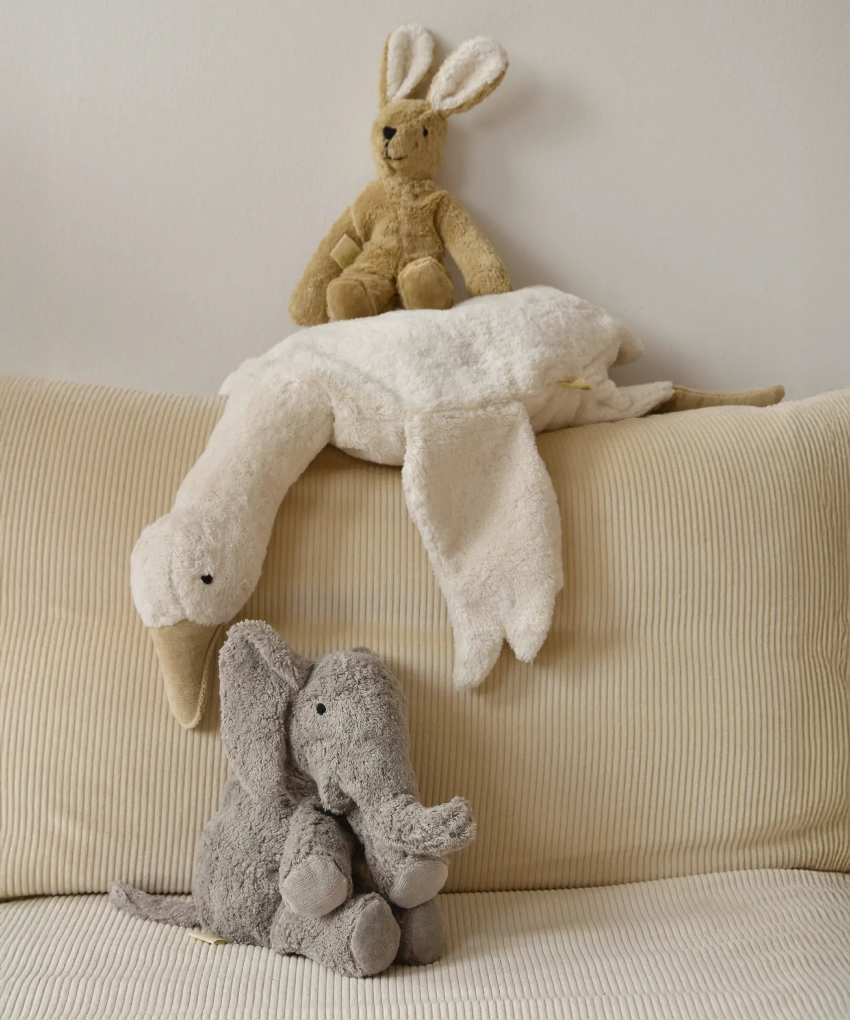 A large Senger elephant cuddly toy with a white goose and beige rabbit soft toy lying above on a corduroy cushion
