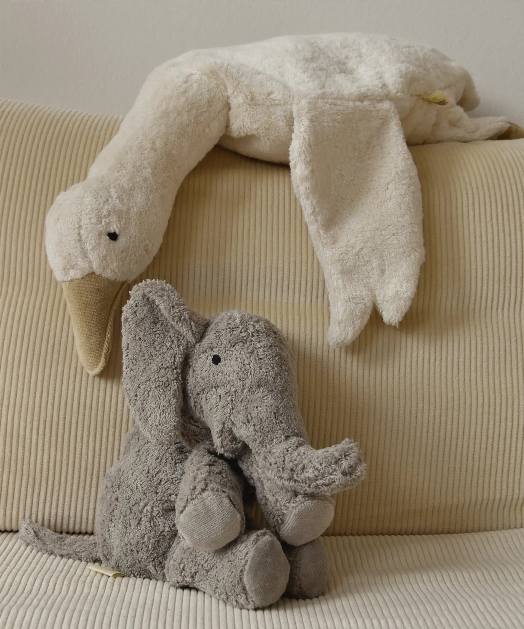 A large Senger elephant cuddly toy with a white goose soft toy lying above on a corduroy cushion