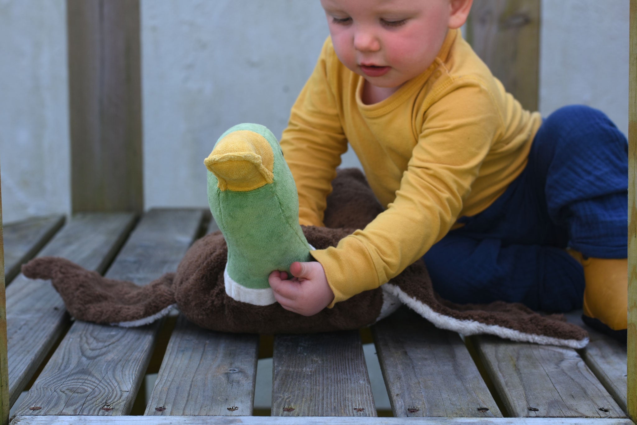 A child sitting on wooden plank holding up the Senger Large Cuddly Drake Duck soft toy's neck.