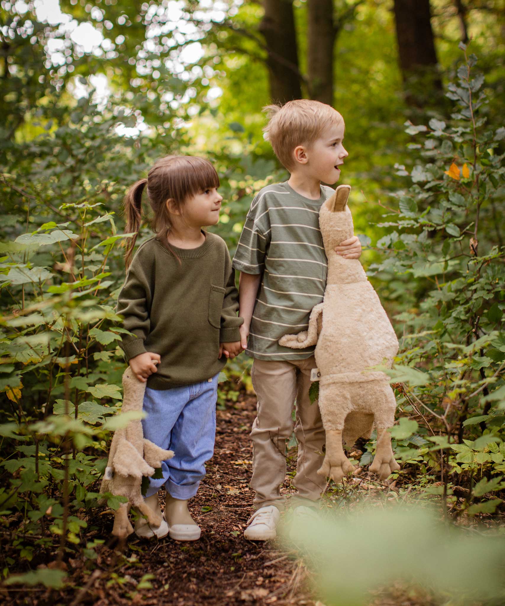 Two children holding hands walking in a forest, one is holding a Senger Large Cuddly Goose organic soft toy in a creamy Linen colour. The other child has the smaller version of the goose. These soft toys are created from the highest quality, sustainable and traceable natural materials. Senger cuddly animal toys and soft dolls are handmade in Germany with love and attention to detail and we are proud Senger stockists here at Babipur. 