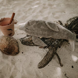 The Senger large organic cotton stuffed dolphin toy, posed on a piece of driftwood on a sandy beach