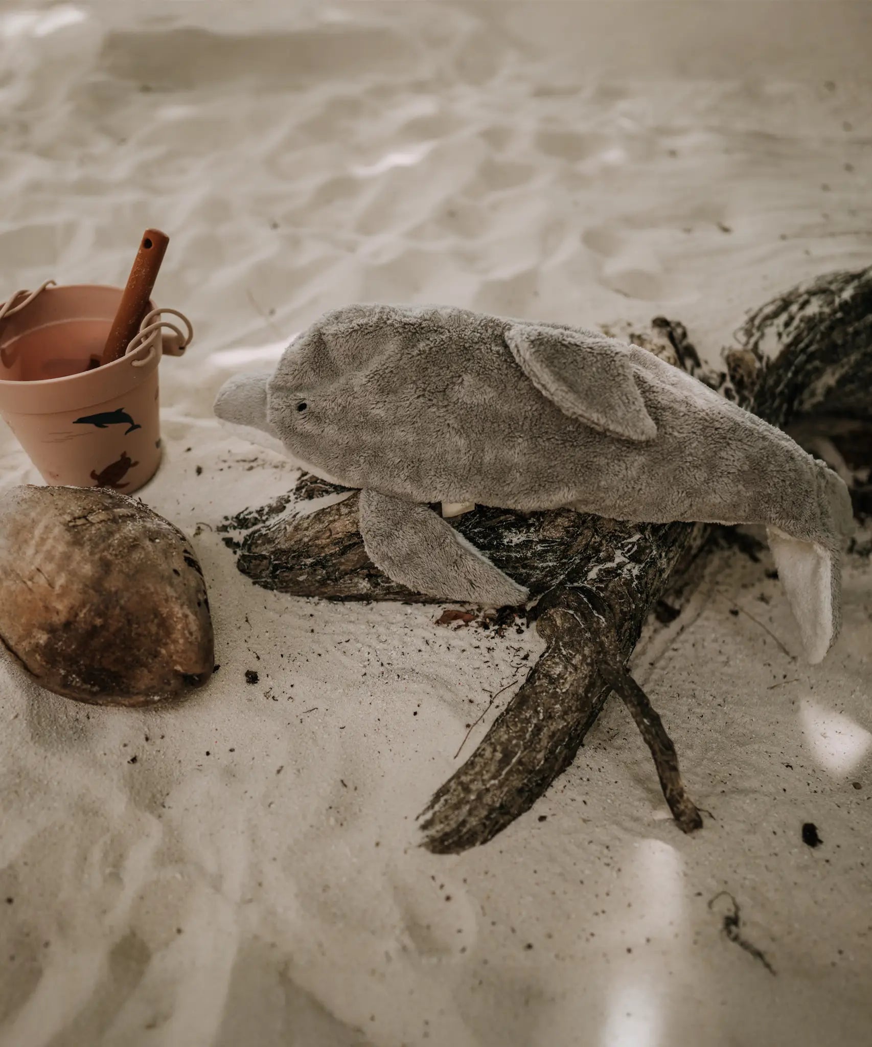 The Senger large organic cotton stuffed dolphin toy, posed on a piece of driftwood on a sandy beach