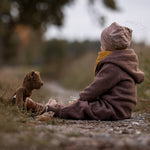 A child sitting on a gravelled surface outdoors with the Senger Large Floppy Brown Bear. 