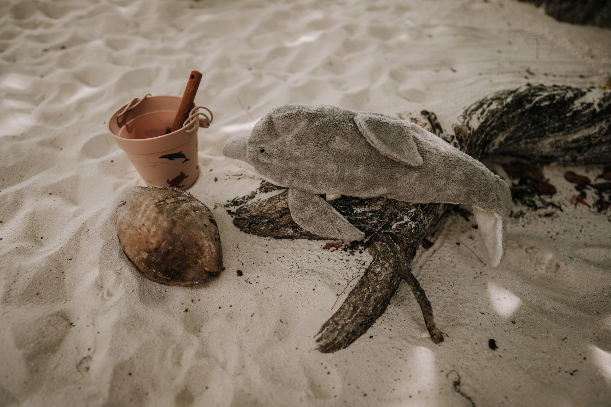 The Senger large organic cotton stuffed dolphin toy, posed on a piece of driftwood on a sandy beach