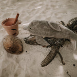 The Senger large organic cotton stuffed dolphin toy, posed on a piece of driftwood on a sandy beach